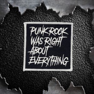 May include: A black square patch with white embroidered text that reads "PUNK ROCK WAS RIGHT ABOUT EVERYTHING." The patch has a white border and is set against a textured, dark background.