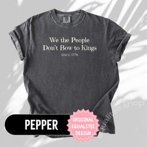 K&ouml;nnte beinhalten: Ein dunkelgraues T-Shirt mit dem wei&szlig;en Text "We the People Don't Bow to Kings SINCE 1776". Das Shirt hat hochgekrempelte &Auml;rmel und einen Rundhalsausschnitt. Das Wort "PEPPER" steht in einem schwarzen Rechteck, und ein rosa Stern sagt "ORIGINAL EQUALITEE DESIGN".
