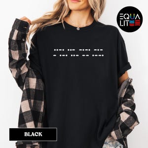 May include: Black t-shirt with the word "EQUAL" written in braille on the front. The shirt is being modeled by a person wearing a plaid shirt.