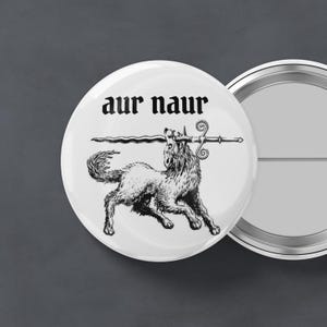 May include: White button with a black and white illustration of a wolf-like creature carrying a sword. The text "aur naur" is printed above the illustration in a gothic font. A second button is visible in the background.