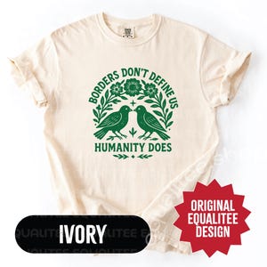 May include: Ivory-colored t-shirt with a green graphic featuring two birds, flowers, and the text "Borders Don't Define Us, Humanity Does." The shirt has a relaxed fit and rolled sleeves. The design is an original Equalitee design.