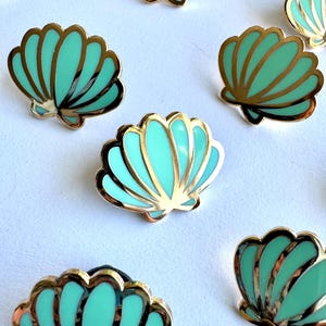 May include: Several teal and gold seashell-shaped enamel pins. The pins have a gold outline and a teal interior. The pins are scattered across a white surface. The pins are a decorative accessory.