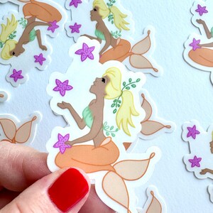 May include: A die-cut sticker featuring a mermaid with blonde hair in a ponytail, a light green top, and an orange tail. The mermaid is surrounded by small purple starfish and green leafy sprigs. The design is printed on a white background.