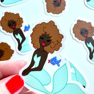 May include: A die-cut sticker featuring a mermaid with dark skin, voluminous brown curly hair, and a bright red lip. The mermaid has a light blue tail and is accompanied by a small blue fish with red lips. The design is repeated across the image.