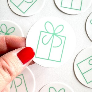 May include: A collection of circular white stickers featuring a simple line drawing of a gift box with a bow in a light mint green color. The stickers are arranged in a group, with one sticker being held up to display the graphic design clearly.