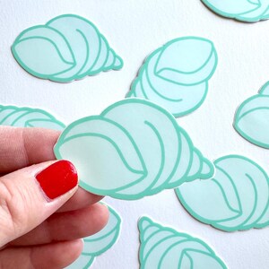 May include: A collection of die-cut stickers shaped like sea shells. Each sticker features a light mint green color with a darker mint green outline, depicting the spiral structure of a shell. The stickers are arranged in a scattered pattern.