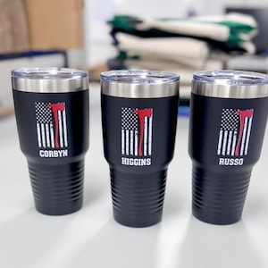 May include: Three black stainless steel tumblers with a distressed American flag design and a red axe graphic. The tumblers are personalized with the names "Corbyn", "Higgins", and "Russo".