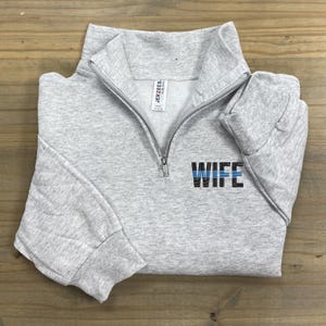 May include: Gray sweatshirt with a zipper and a blue line thin blue line design with the word "WIFE" on the front.
