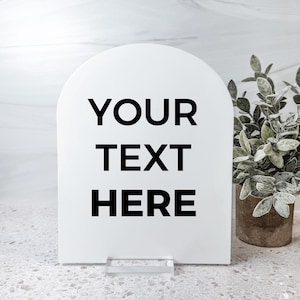 May include: A white arched sign with the text "YOUR TEXT HERE" in black. The sign is supported by a clear acrylic base. A potted plant with green leaves is visible in the background.