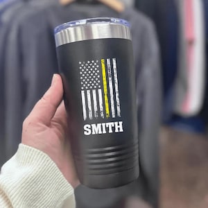 May include: Black insulated tumbler with a silver rim and lid. The tumbler features a distressed American flag design with a yellow stripe and the name "SMITH" in white block letters. The tumbler is cylindrical and appears to be made of stainless steel.