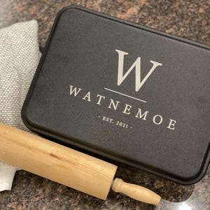 May include: Black baking pan with a white monogram "W" and the text "WATNEMO E - EST. 2021 -" on the front. A wooden rolling pin is in the foreground.