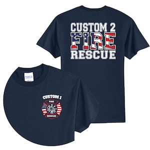 May include: A navy blue t-shirt featuring a front left chest graphic of a red and white fire rescue emblem with two American flags and the text CUSTOM 1 FIRE RESCUE. The back displays the text CUSTOM 2 FIRE RESCUE with an American flag pattern.