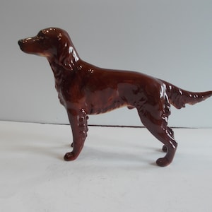 May include: A glossy, brown ceramic dog figurine in a standing pose. The dog has a long, slender body, with its tail slightly raised. The figurine is a decorative item, likely for display.