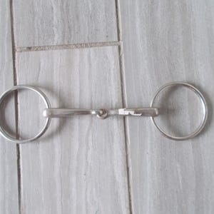 May include: A silver horse bit with two rings. The bit has a jointed mouthpiece.