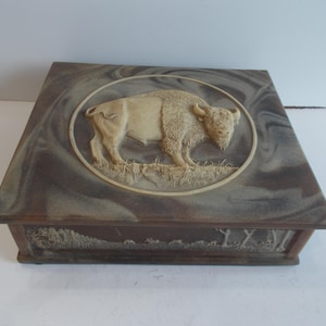 May include: A square, wooden box with a decorative bison relief on the lid. The bison is cream-colored and set within an oval frame. The box has a marbled, brown and gray finish. The sides of the box feature a landscape design.