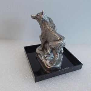 May include: A silver-colored wolf sculpture perched on a rock, set on a black square base. The wolf is depicted in a seated position, looking upwards. The sculpture has detailed fur and a realistic appearance.