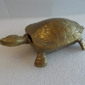May include: A brass turtle figurine with a hinged shell that opens to reveal a hollow interior. The metal surface features a polished gold-toned finish and engraved geometric patterns on the shell, head, and legs, showing a realistic shape.