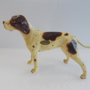 Puede incluir: Una figurita de cerámica de un perro manchado de marrón y blanco. El perro está de pie sobre sus cuatro patas con la cola enroscada. La figurita tiene una etiqueta marrón y dorada en un lado que dice "Royal Doulton  Inglaterra".