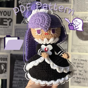 May include: Crocheted amigurumi doll with purple hair, a black dress with white lace trim, and a white flower. The doll has a brown face with purple eyes and a red mouth.