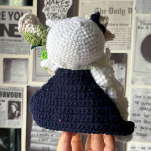 May include: A crocheted doll with white hair and a dark blue dress. The doll is holding a bouquet of crocheted flowers.