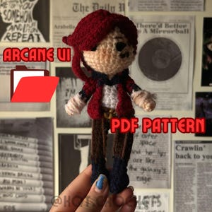 May include: Crochet pattern for a red and brown amigurumi doll with a red hat and a black shirt. The doll is holding a red folder with the text "ARCANE UI" on it. The text "PDF PATTERN" is also visible in the image.
