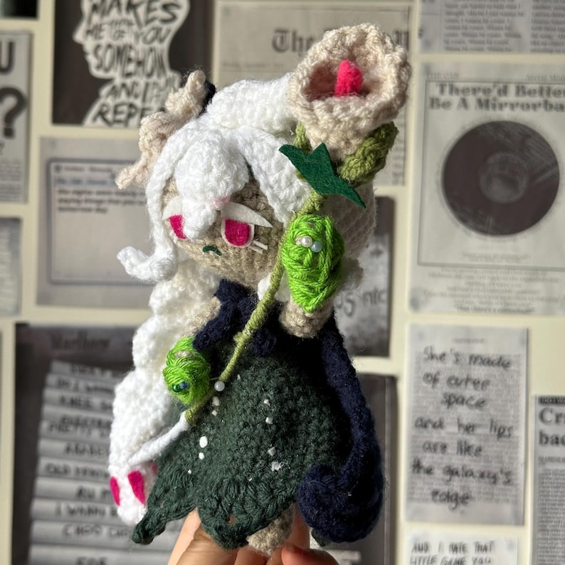 May include: A crocheted plush toy with white hair, a green dress, and a pink flower. The toy is holding a green stem with a pink flower.