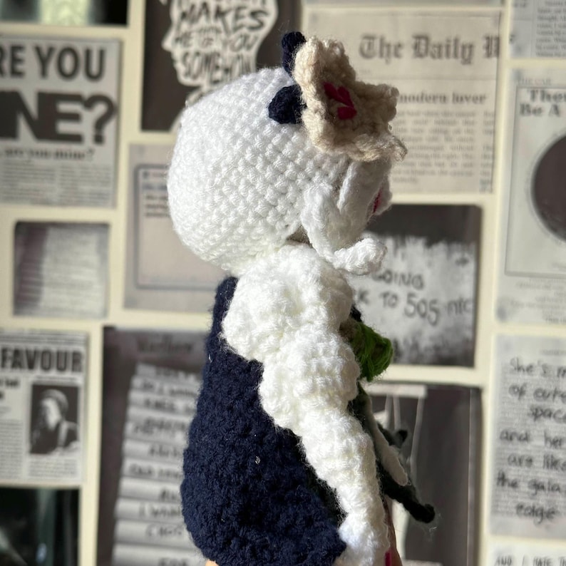 May include: A white crocheted doll wearing a black and white striped dress and a white crocheted hat. The doll has a pink flower on its head and is holding a green object in its hand.