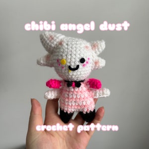 May include: A handmade crochet doll of a character with white fur, pink accents, and black shoes. The doll has a yellow eye, a black smile, and a black bow tie. The text "chibi angel dust" and "crochet pattern" are displayed in pink.