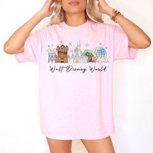 May include: A light pink short-sleeve t-shirt featuring a colorful graphic of the Walt Disney World skyline, including the castle, iconic park structures, and small stars. The text Walt Disney World is printed in black script below the illustration.