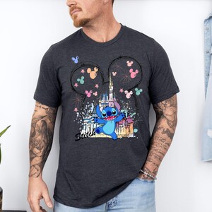 May include: Dark gray t-shirt featuring a colorful graphic of Stitch from Lilo & Stitch in front of a castle, with Mickey Mouse ears and fireworks. The word "Stitch" is written in a script font. The shirt is short-sleeved.
