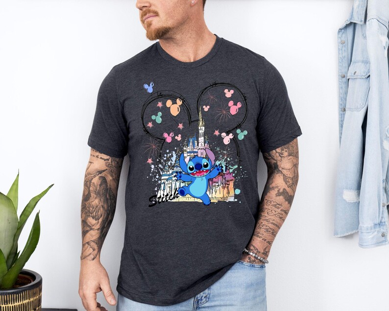 May include: Dark gray t-shirt featuring a colorful graphic of Stitch from Lilo & Stitch in front of a castle, with Mickey Mouse ears and fireworks. The word "Stitch" is written in a script font. The shirt is short-sleeved.