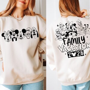 Peut inclure: Sweat-shirt cr&egrave;me avec un motif noir et blanc de personnages Disney. L'avant pr&eacute;sente des visages de personnages, tandis que l'arri&egrave;re affiche "Family Vacation 2026" avec d'autres personnages.