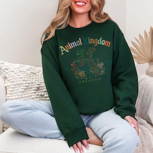 K&ouml;nnte beinhalten: Ein dunkelgr&uuml;nes Sweatshirt mit dem Schriftzug "Animal Kingdom" in Regenbogenfarben. Darunter befindet sich eine Tiergrafik und das Wort "Orlando". Das Sweatshirt hat lange &Auml;rmel und einen Rundhalsausschnitt.