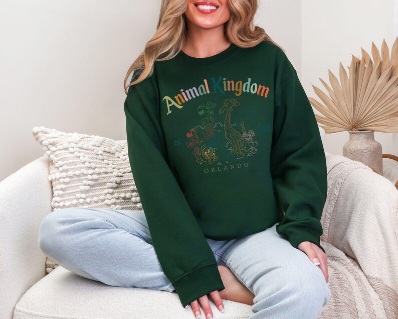 K&ouml;nnte beinhalten: Ein dunkelgr&uuml;nes Sweatshirt mit dem Schriftzug "Animal Kingdom" in Regenbogenfarben. Darunter befindet sich eine Tiergrafik und das Wort "Orlando". Das Sweatshirt hat lange &Auml;rmel und einen Rundhalsausschnitt.