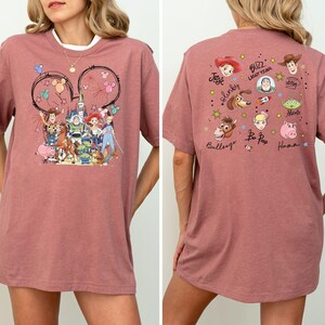 Op de afbeelding: Een stoffig roze t-shirt met een kleurrijke afbeelding van Toy Story-personages. Het ontwerp aan de voorkant bevat Woody, Buzz Lightyear en andere personages. De achterkant van de shirt heeft extra karakterillustraties en namen.