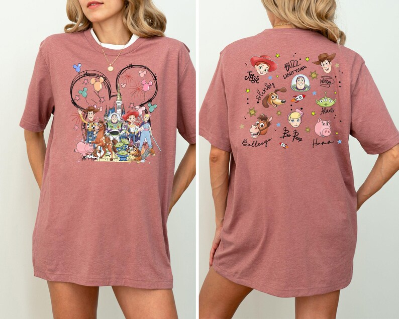 Op de afbeelding: Een stoffig roze t-shirt met een kleurrijke afbeelding van Toy Story-personages. Het ontwerp aan de voorkant bevat Woody, Buzz Lightyear en andere personages. De achterkant van de shirt heeft extra karakterillustraties en namen.
