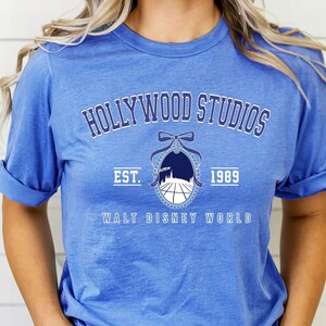 Puede incluir: Camiseta azul con mangas enrolladas que muestra el texto "HOLLYWOOD STUDIOS" sobre un emblema y el texto "EST. 1989 WALT DISNEY WORLD". El emblema tiene un lazo y una silueta de paisaje urbano.