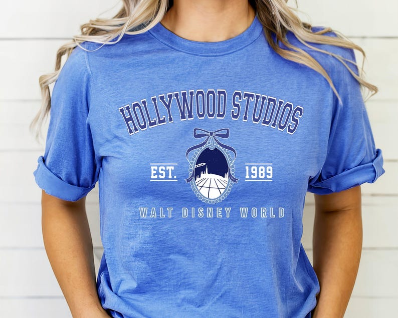 Puede incluir: Camiseta azul con mangas enrolladas que muestra el texto "HOLLYWOOD STUDIOS" sobre un emblema y el texto "EST. 1989 WALT DISNEY WORLD". El emblema tiene un lazo y una silueta de paisaje urbano.