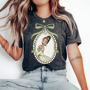 May include: A dark gray t-shirt with a green and white illustration of a princess in a pearl frame. The princess has brown skin, brown eyes, and a crown. The illustration is surrounded by a green bow.