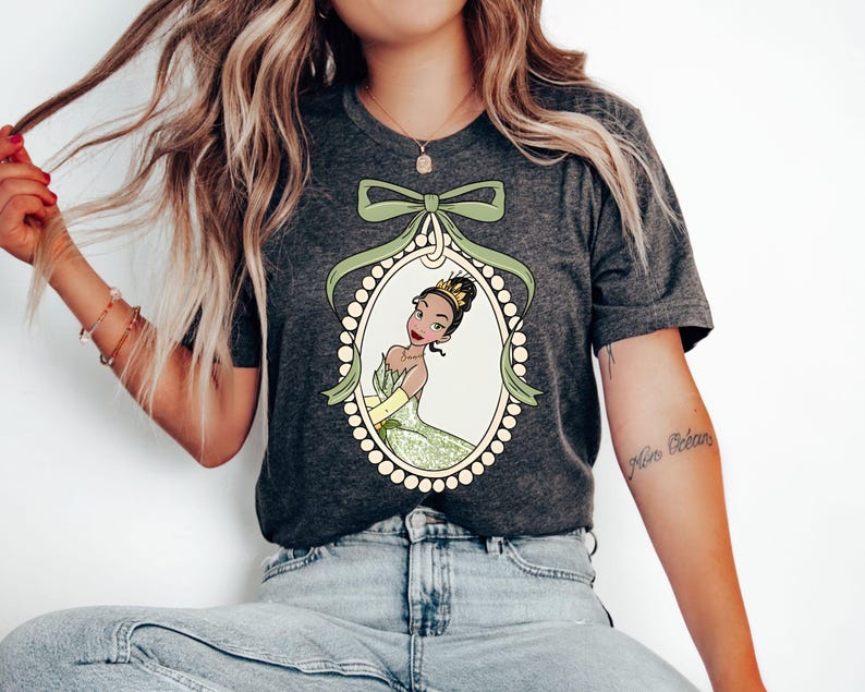 May include: A dark gray t-shirt with a green and white illustration of a princess in a pearl frame. The princess has brown skin, brown eyes, and a crown. The illustration is surrounded by a green bow.
