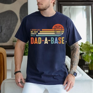 Könnte beinhalten: Marineblaues T-Shirt mit dem Text "I keep all my dad jokes in a Dad-a-base" in einem Retro-Design. Die Grafik enthält einen Sonnenuntergang und eine Baseball-Homeplate. Das Shirt besteht aus einem weichen, bequemen Material.