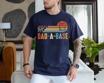 Op de afbeelding: Marineblauw T-shirt met de tekst "I keep all my dad jokes in a Dad-a-base" in een retro-ontwerp. De afbeelding bevat een zonsondergang en een honkbalplaat. Het shirt is gemaakt van een zacht, comfortabel materiaal.