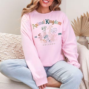 K&ouml;nnte beinhalten: Ein hellrosa Sweatshirt mit den Worten "Animal Kingdom" in Regenbogenfarben und einer Grafik von Zeichentricktieren. Das Sweatshirt hat auch die Worte "est. 1998 Orlando".