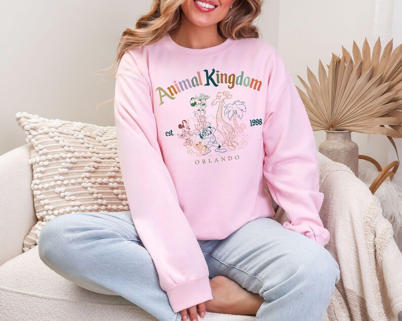 K&ouml;nnte beinhalten: Ein hellrosa Sweatshirt mit den Worten "Animal Kingdom" in Regenbogenfarben und einer Grafik von Zeichentricktieren. Das Sweatshirt hat auch die Worte "est. 1998 Orlando".