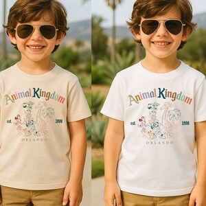K&ouml;nnte beinhalten: Zwei kurz&auml;rmelige T-Shirts, eines cremefarbenes und eines wei&szlig;es, zeigen den Text "Animal Kingdom" in Regenbogenfarben, mit einer Grafik von Zeichentieren und dem Text "Orlando 1998".