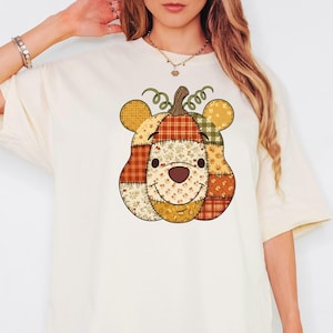 May include: A cream-coloured t-shirt with a patchwork pumpkin design featuring a bear face. The pumpkin is made of various patterned fabrics in orange, yellow, and brown, with a smiling face and a green stem. The design is centered on the shirt.