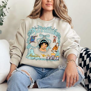 Pu&ograve; includere: Felpa beige con un'illustrazione turchese e bianca della principessa Jasmine di Aladdin. Il design include la parola "Jasmine" in corsivo turchese, un ritratto di Jasmine e altri personaggi del film.