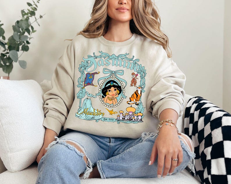 Pu&ograve; includere: Felpa beige con un'illustrazione turchese e bianca della principessa Jasmine di Aladdin. Il design include la parola "Jasmine" in corsivo turchese, un ritratto di Jasmine e altri personaggi del film.