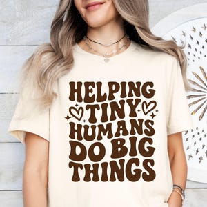 May include: Cream-colored t-shirt with the text "HELPING TINY HUMANS DO BIG THINGS" in brown, retro-style lettering. The design includes heart and star accents. The shirt is a casual, everyday wear item.