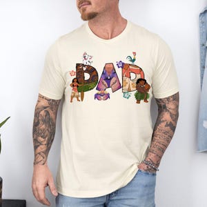 K&ouml;nnte beinhalten: Ein cremefarbenes T-Shirt mit einem Aufdruck des Wortes "DAD", das aus Figuren aus dem Film Vaiana besteht. Die Figuren sind Maui, Vaiana, das Schwein Pua, der Hahn Hei Hei und der Fischhaken des Halbgottes Maui.